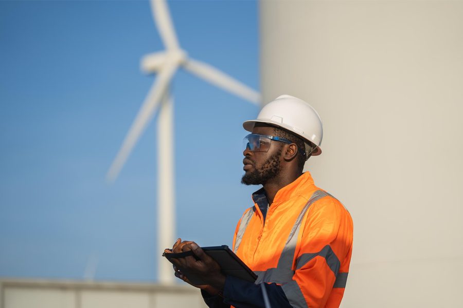 An engineer in hi-vis and a hard hat reviewing data on a tablet beside a wind turbine