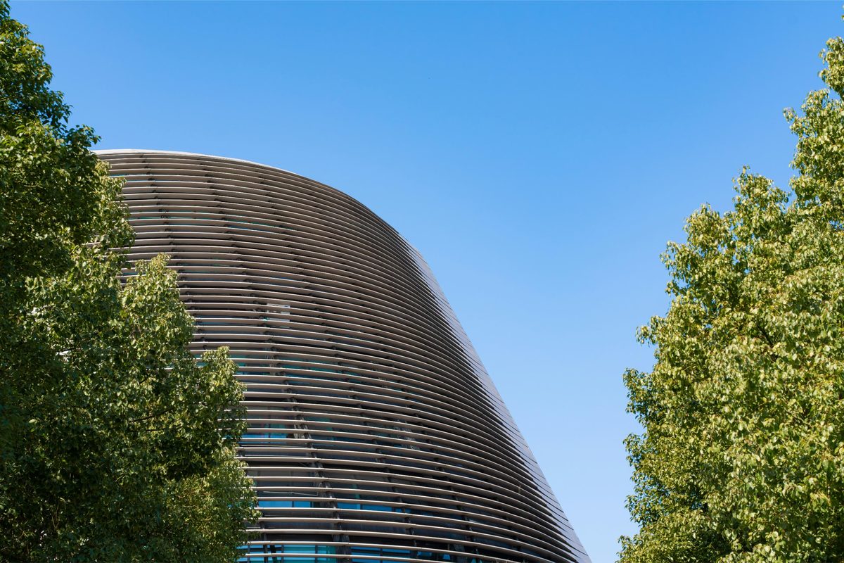 A curved building clad in horizontal fins, framed by trees against a blue sky